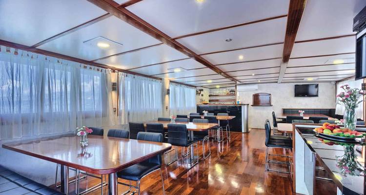 Yacht dining salon with polished wood floors, tables, and large curtained windows.