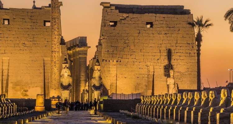 Statues and avenue of sphinxes lead toward towering pylons of Luxor Temple at dusk.