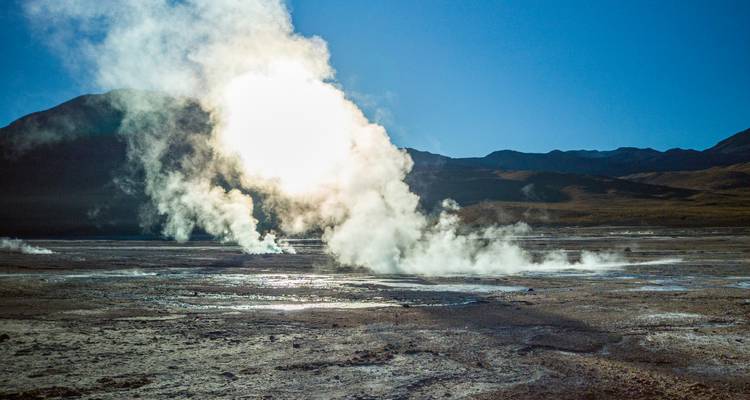 Steam rises from active geysers on an arid plateau with distant Andean peaks under a clear blue sky.