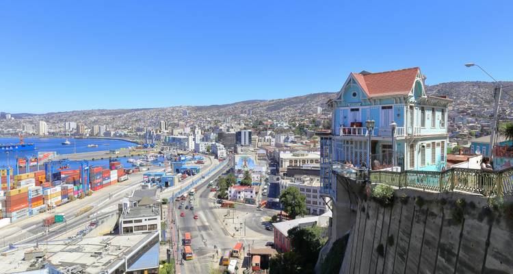 Colorful cliff-top Victorian house overlooks the busy Valparaiso port and city sprawl on a bright day.