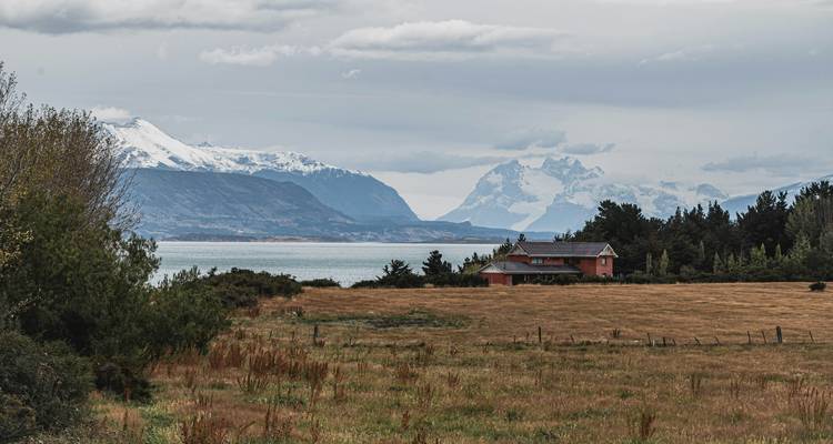 Rural farmhouse on grassy plain overlooks a fjord with snowy Patagonian peaks hidden in cloud.