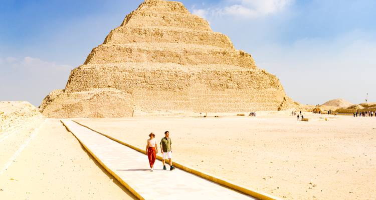 Ein Paar spaziert auf einem langen Weg zur Stufenpyramide von Saqqara unter diesigem, blauem Himmel.