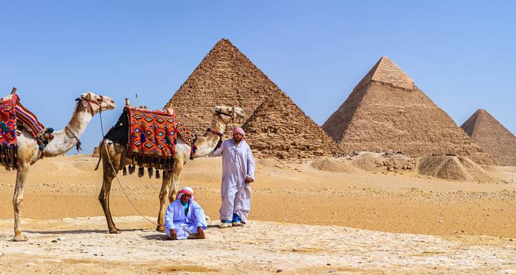 Zwei Kamele und ihre Führer vor den ikonischen Pyramiden von Giza unter klarem Himmel.