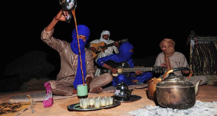 Tuareg-muzikanten en gastheren die thee zetten naast een kampvuur in de woestijnnacht.