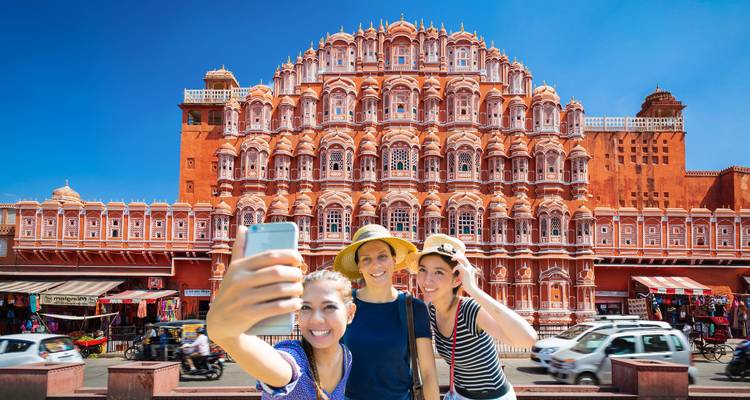 Drei lächelnde Touristen, die ein Selfie vor Jaipurs verziertem rosa Hawa Mahal unter einem strahlend blauen Himmel machen.