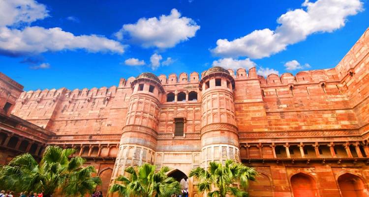 Red sandstone walls and massive twin gates of Agra Fort glowing under a vibrant blue sky with scattered clouds.