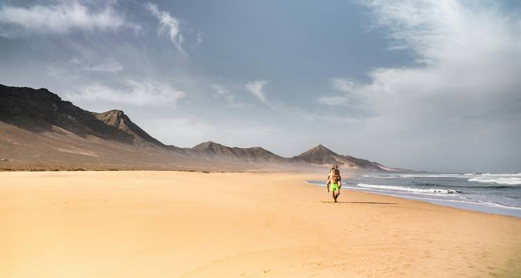 Breiter leerer Sandstrand mit einzelnem Surfer, der ein Brett zu den weit entfernten Wellen und Bergen trägt