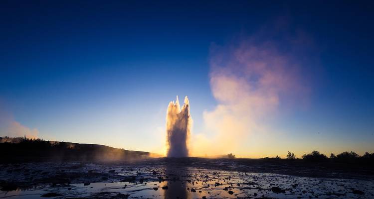 Ein mächtiger Geysir bricht hoch in den Himmel gegen ein tiefblaues Morgenlicht aus, während Dampf aus dem geothermischen Feld aufsteigt.