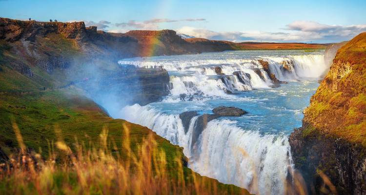 Ein dramatischer, mehrstöckiger Wasserfall, der sich durch grüne Klippen schneidet und einen Regenbogen in der Gischt bildet.