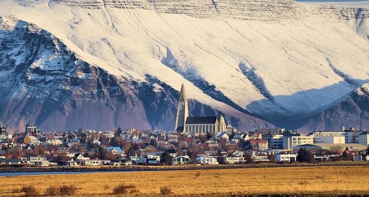 Reykjaviker Stadtbild, das von der Hallgrimskirkja-Kirche dominiert wird und von schneebedeckten Bergen in warmem Licht eingerahmt ist.