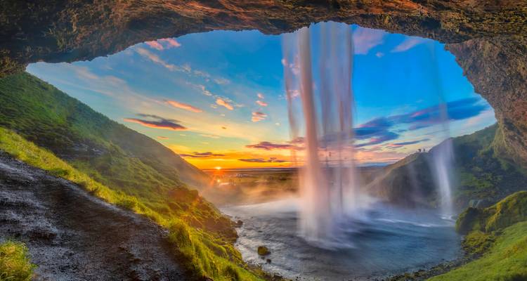 Aussicht von hinter einem imposanten Wasserfall, der in ein grünes Tal bei einem farbenfrohen Sonnenuntergang hinabstürzt.