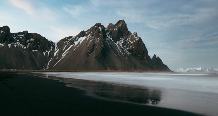Scharfkantige schwarze Vulkangipfel des Vestrahorn erheben sich über einen glatten schwarzen Sandstrand unter weichem Licht.