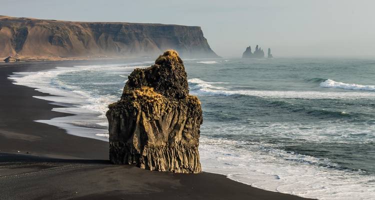 Schwarzer Vulkanstrand mit moosbedecktem Felsenpfeiler und entfernten Reynisdrangar-Säulen, die sich aus nebligen Wellen erheben.