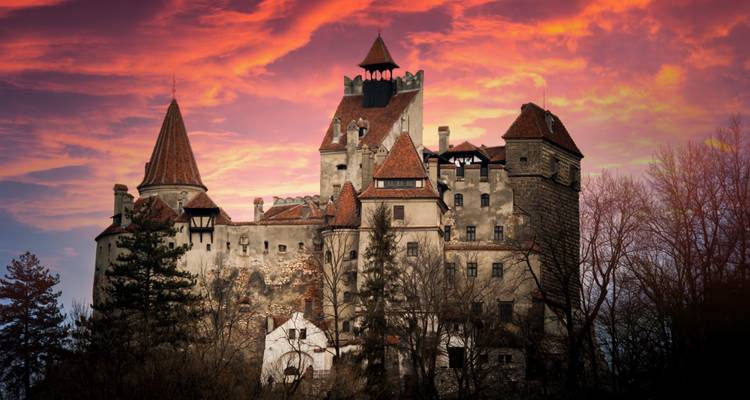 Le château de Bran se détachant en silhouette contre un ciel de coucher de soleil dramatique aux teintes rouges et pourpres.