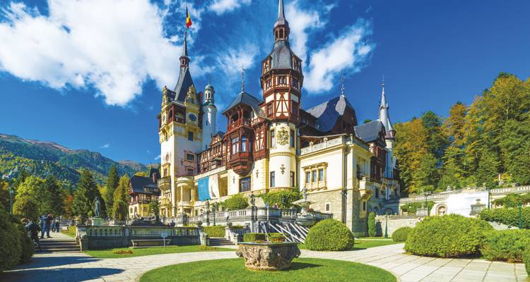 Élégant château de Peles avec ses tourelles ornées et ses jardins impeccablement entretenus encadrés par des montagnes boisées par une journée dégagée.