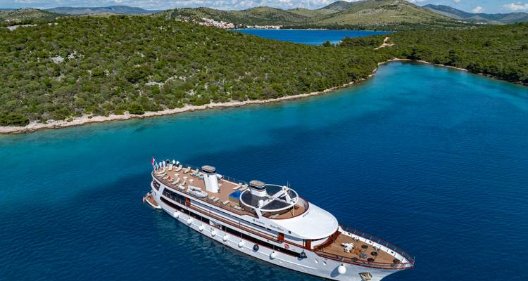Vista aérea de un elegante yate de crucero blanco anclado en aguas azules intensas junto a una isla croata cubierta de bosques.