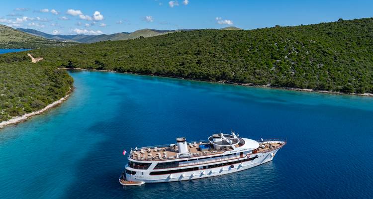 Imagen de dron de un yate de crucero deslizándose por una ensenada turquesa con colinas verdes y cielo azul.