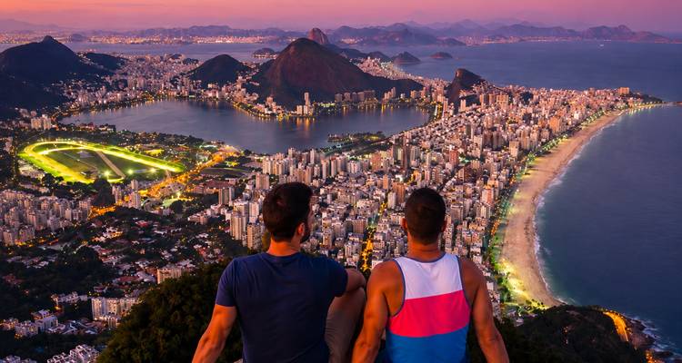 Dos viajeros se sientan muy arriba de Rio de Janeiro, mirando playas, lagunas y montanas al atardecer.