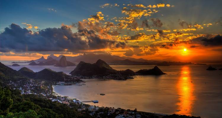 Un atardecer ardiente se refleja en la Bahía de Guanabara con el Pan de Azúcar y picos distantes bajo nubes dramáticas.