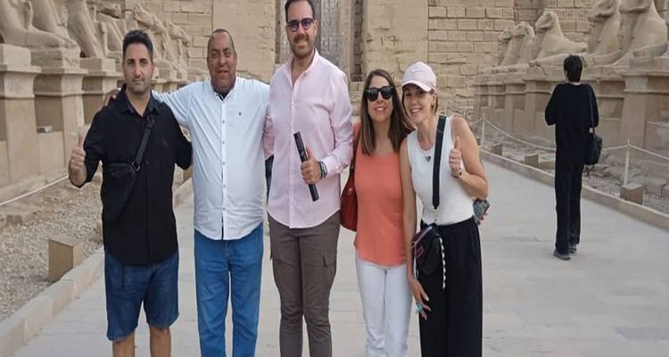 Five travellers pose with thumbs up on a pathway lined with sphinx statues at an ancient site.
