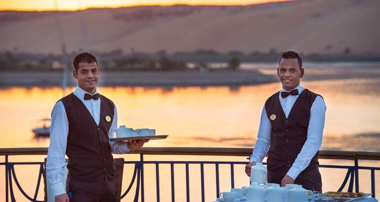 Two smartly dressed waiters holding trays of tea cups on a Nile cruise deck at sunset with golden dunes beyond.