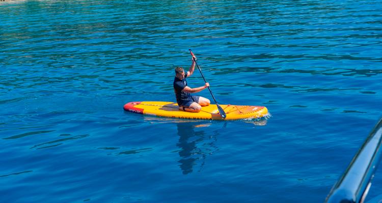 Person kneeling on stand-up paddleboard gliding over clear blue water beside ship.