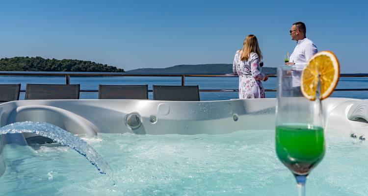 Jacuzzi on deck with colourful cocktail in foreground and couple conversing beside sea backdrop.