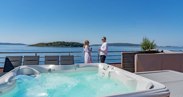 Wide shot of deck jacuzzi with couple holding drinks against calm Adriatic islands.