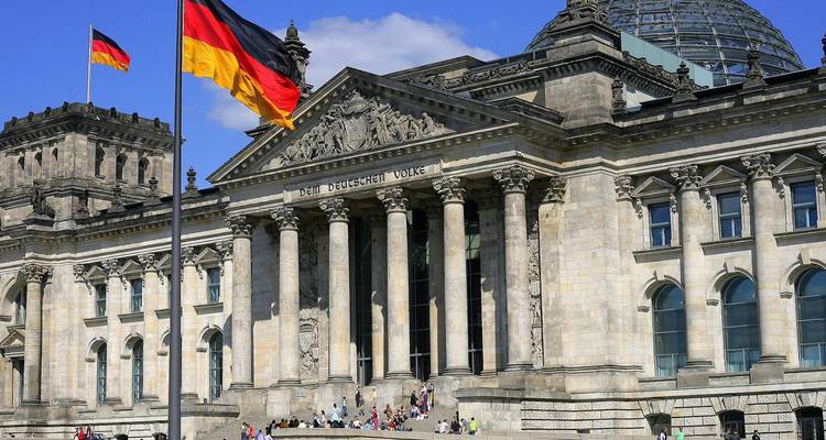 Le drapeau allemand flotte devant le bâtiment néoclassique du Reichstag de Berlin sous un ciel dégagé.