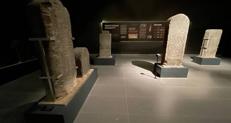Dimly lit museum gallery displaying tall ancient stone pillars and inscribed stelae.
