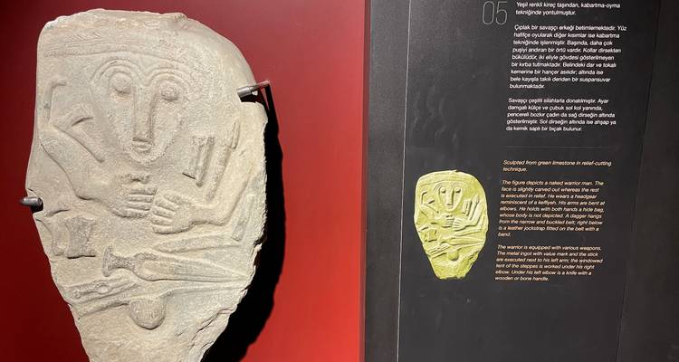 Close-up museum display of a carved stone mask alongside explanatory text on a red and black panel.