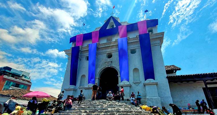 White colonial church draped in purple festival banners with locals gathered on stone steps under a bright sky in Chichicastenango