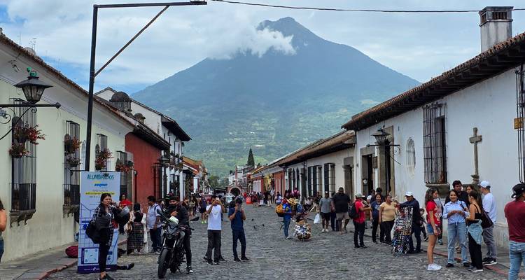 Cobbled street in Antigua leading toward a cloud-topped volcano with people strolling and colonial buildings on both sides