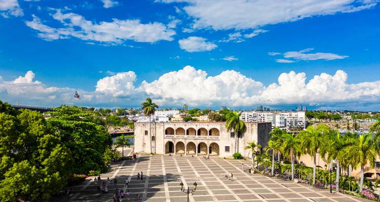 Colonial plaza in Santo Domingo with historic stone building, palm trees and locals strolling across the square.