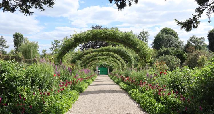 Allée de jardin bordée de fleurs encadrée par des treillages verts en arche menant vers un portail par une journée ensoleillée.