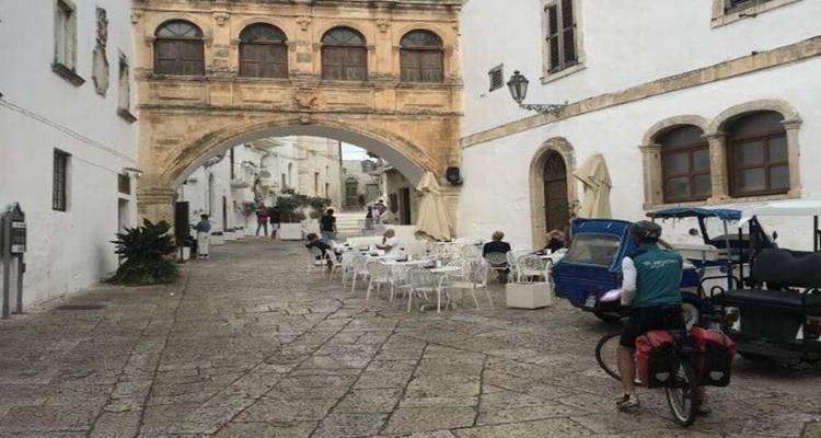 Les cyclistes passent sous une arche en pierre historique dans une rue étroite aux murs blanchis à la chaux avec des tables de café en terrasse.