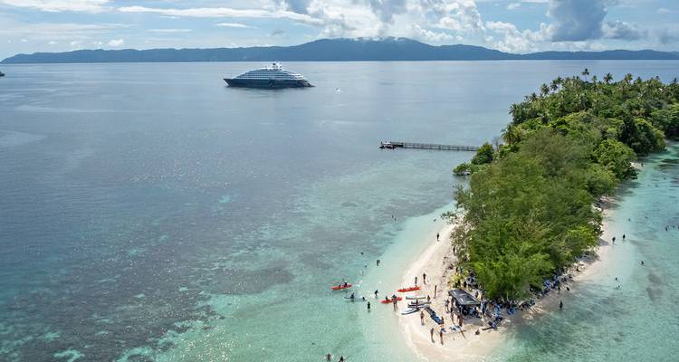 Yate de crucero de lujo anclado frente a una pequeña isla tropical con playa de arena blanca y kayakistas.