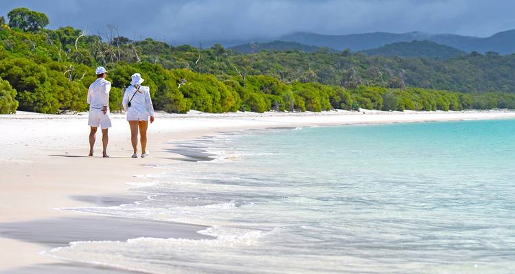 Couple vêtu de blanc marchant le long d'une plage de sable blanc immaculé avec de l'eau turquoise et un arrière-plan forestier.