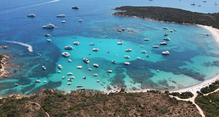 Luftaufnahme einer türkisfarbenen Bucht mit Yachten und Booten, die in der Nähe eines Sandstrandes und einer bewaldeten Halbinsel vor Anker liegen.