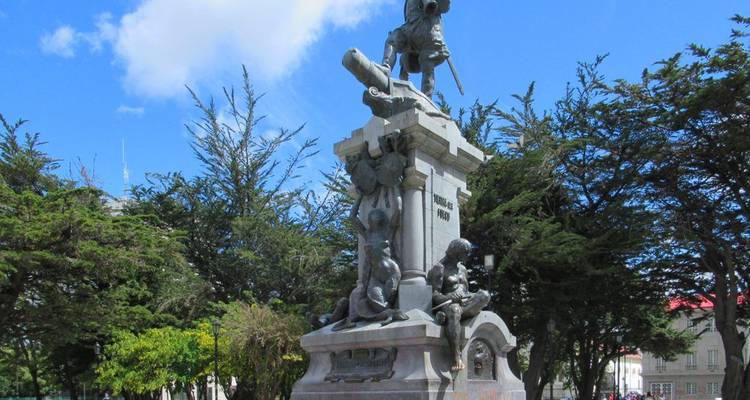 Monument featuring explorer statue and cannon in a tree-filled plaza under a blue sky.