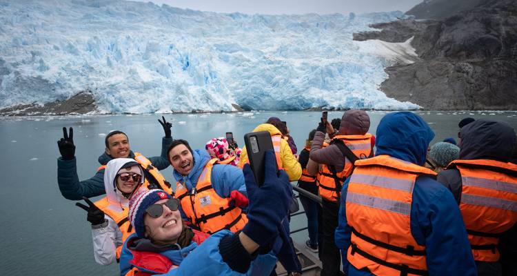 Bundled in orange life-jackets, smiling travellers take selfies on a boat in front of a towering blue glacier.