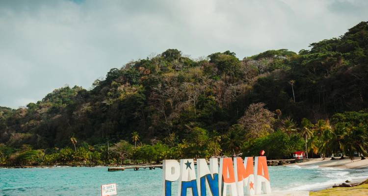 Panneau de plage coloré « PANAMA » situé sur une eau turquoise avec une colline boisée en arrière-plan.