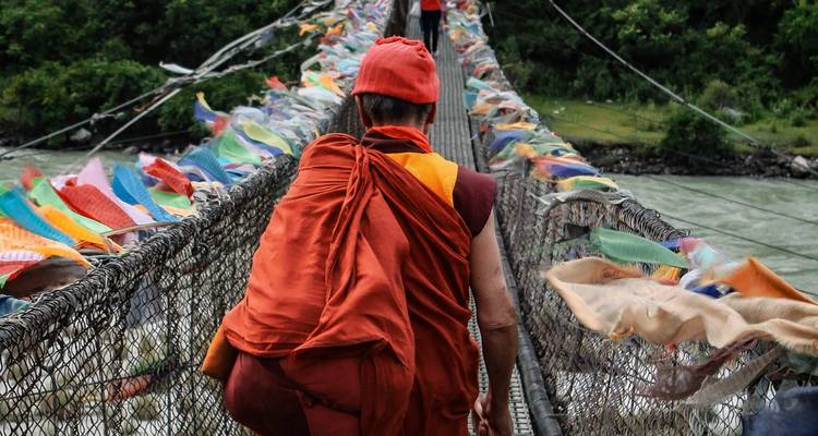 Monk in saffron robe crossing a long suspension bridge draped with colorful prayer flags over a river