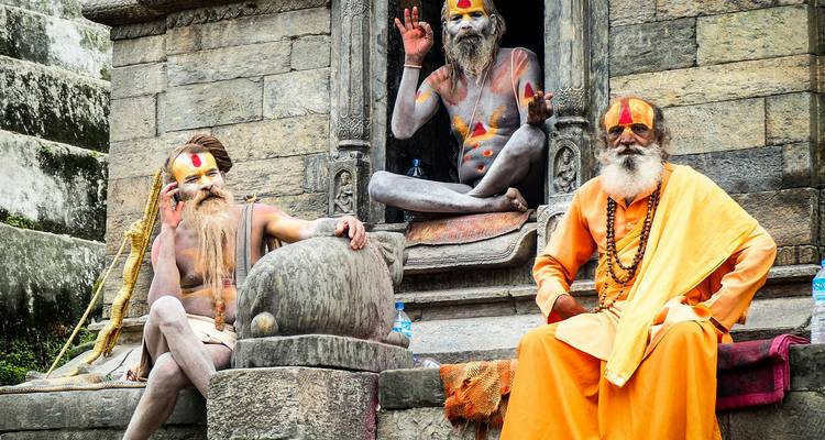 Three colorfully painted sadhus pose and gesture outside a stone temple shrine.
