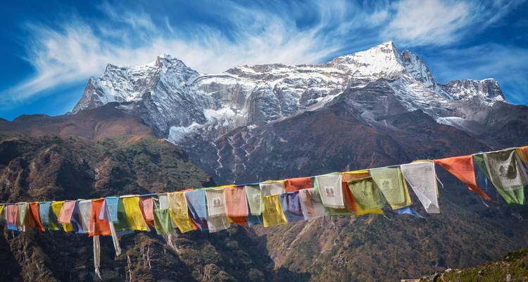 Prayer flags flutter in the foreground with dramatic Himalayan peaks and wispy clouds behind.