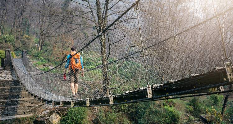 A hiker with an orange backpack crosses a long suspension bridge over a forested valley.