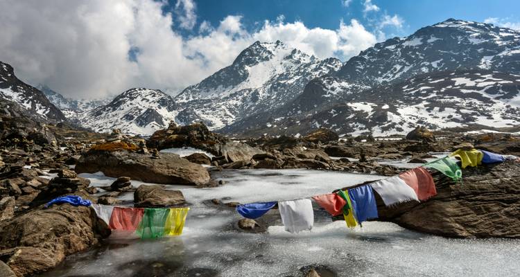 Frozen mountain stream surrounded by snow-dusted rocks and Himalayan peaks with prayer flags stretched across.