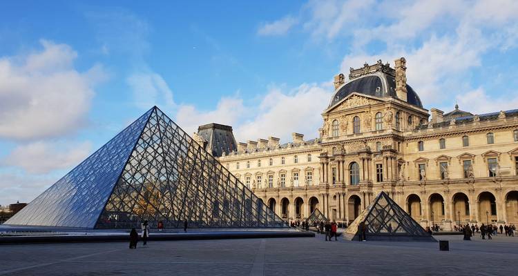 Glass pyramid of the Louvre Museum gleams in front of the historic palace under a bright sky.
