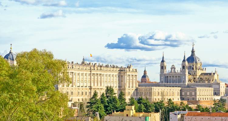 Distant view of Madrid’s Royal Palace and Almudena Cathedral rising above treetops under partly cloudy sky.