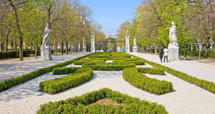 Formal hedged walkway in Madrid’s Retiro Park with statues and strolling visitors on a spring day.